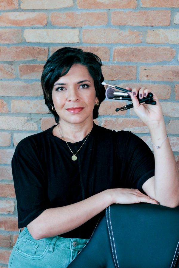 Makeup artist holding brushes with a confident smile against brick wall background.